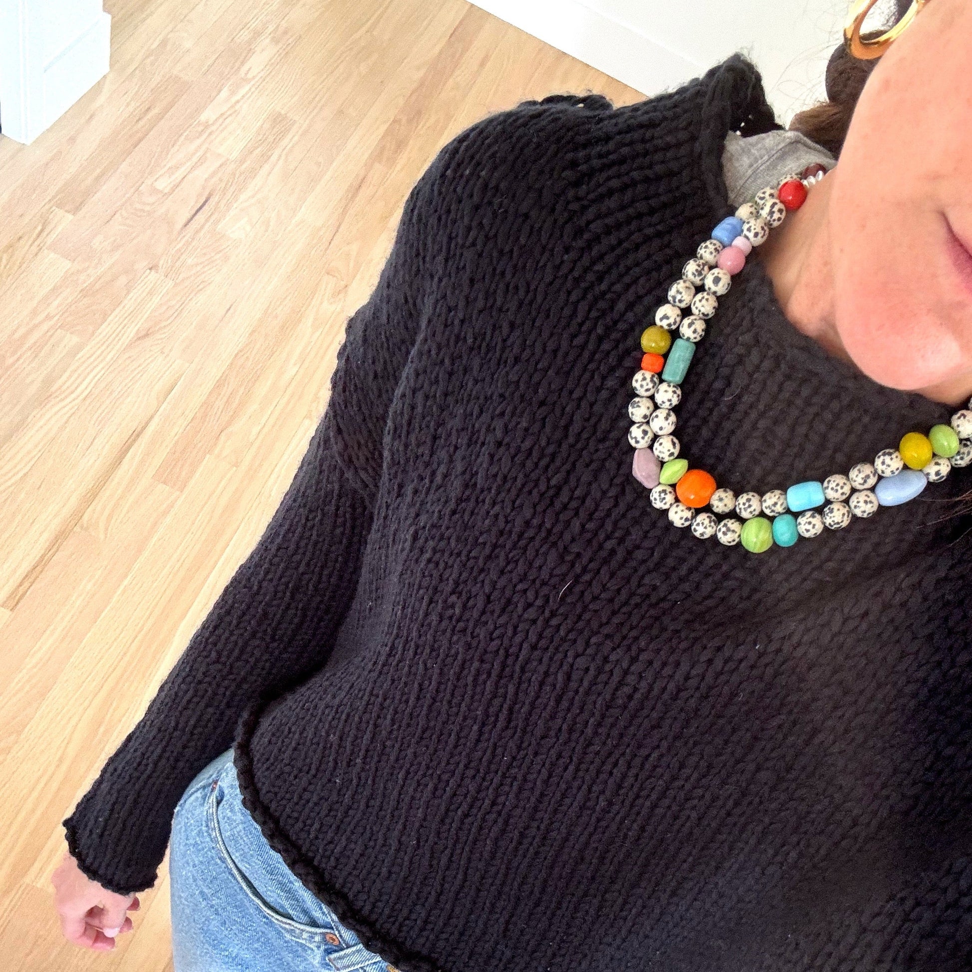 Person wearing a black sweater and colorful beaded necklace on a wooden floor.