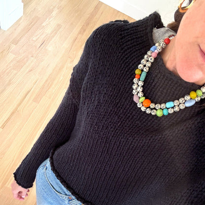Person wearing a black sweater and colorful beaded necklace on a wooden floor.