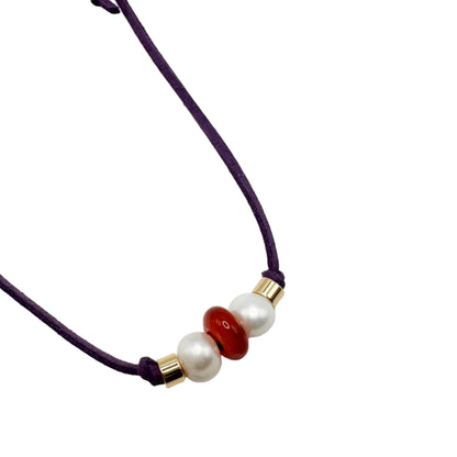 A close-up of a purple cord necklace featuring three central beads: two white pearls and one glossy red bead, accented with small gold spacers, on a plain white background.