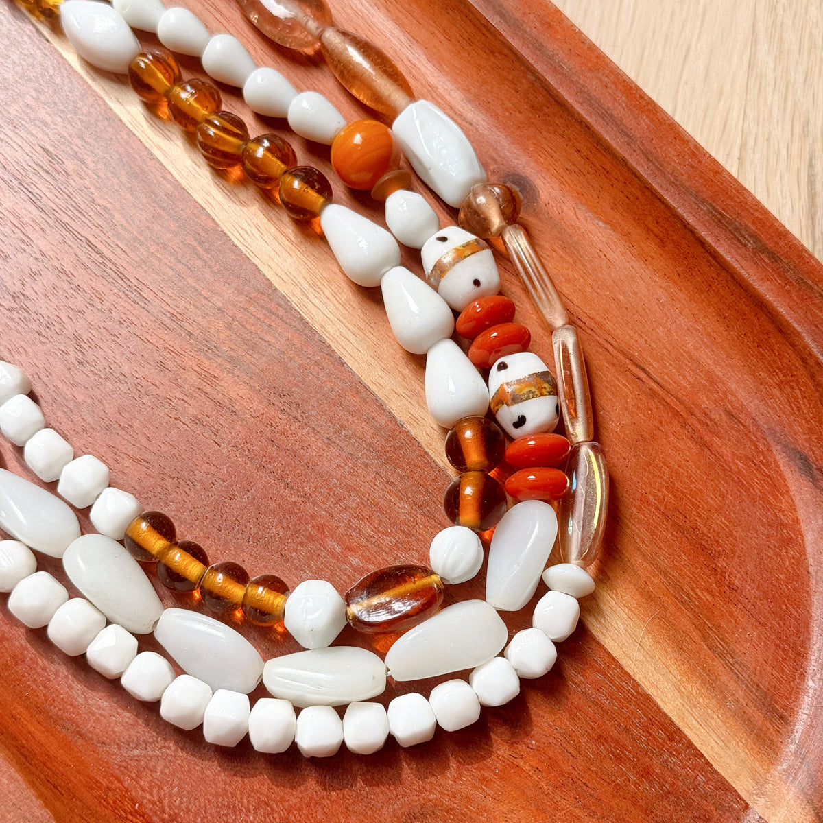 A close-up of a beaded necklace with white stone beads, translucent amber-brown beads, and a decorative red and white patterned bead, arranged on a curved wooden tray.