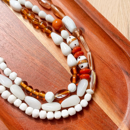 A close-up of a beaded necklace with white stone beads, translucent amber-brown beads, and a decorative red and white patterned bead, arranged on a curved wooden tray.