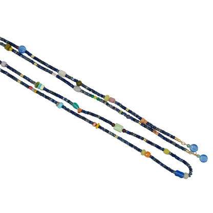 A long beaded necklace laid diagonally on a plain white background, featuring dark blue beads with scattered colorful accents in blue, green, orange, pink, yellow, and white.