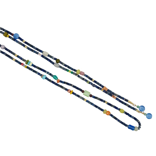 A long beaded necklace laid diagonally on a plain white background, featuring dark blue beads with scattered colorful accents in blue, green, orange, pink, yellow, and white.
