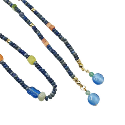 A beaded necklace with dark blue and gray beads, small gold spacer beads, and a drop section featuring a blue cylindrical bead, a light green bead, and two blue oval pendants, displayed on a white background.