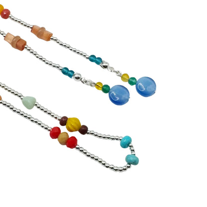 A close-up of two silver beaded necklaces laid flat on a white background. One necklace features translucent orange stone beads, while the other features a large round blue bead, turquoise, yellow, and red beads.