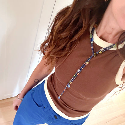 A person wearing a brown ribbed knit top with cream trim, bright blue pants, and a colorful beaded necklace, standing on a light wood floor.